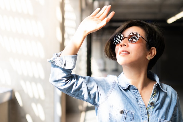 Asian woman wearing 100% UV light eyes protection sunglasses, stand and raise her hand to block out bright glare and sunlight